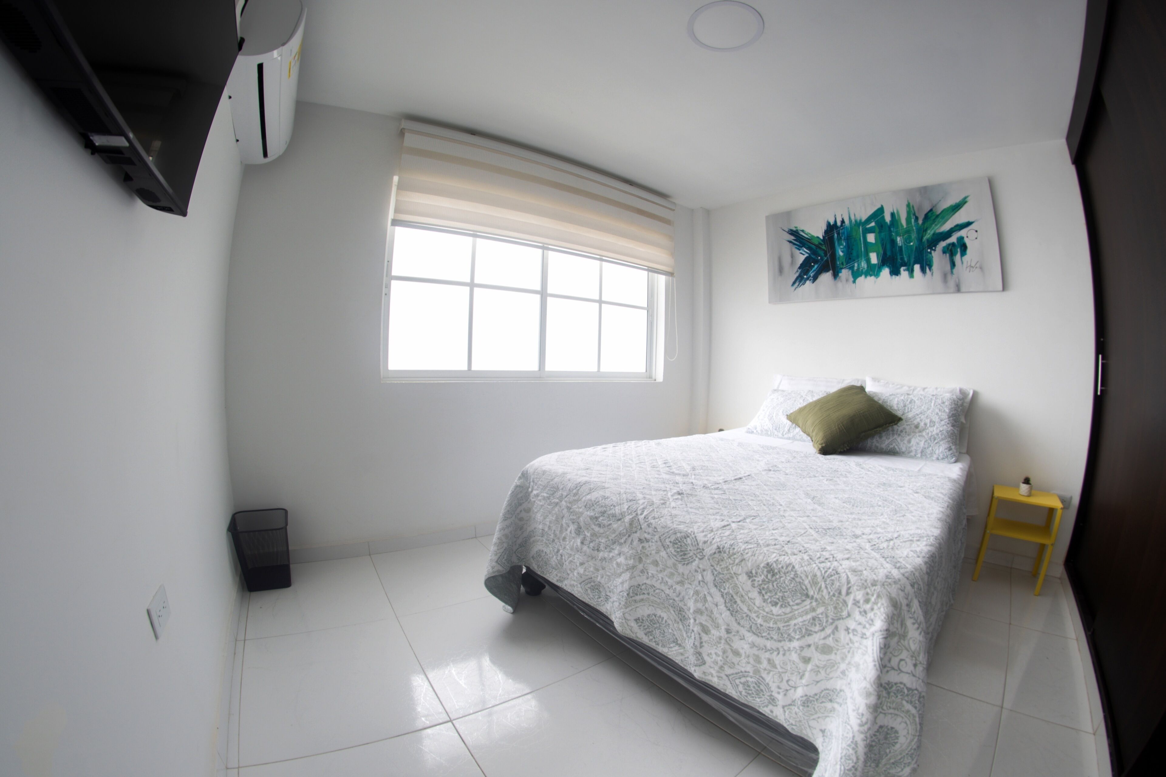 A bedroom with a bed, a window with blinds, a small bedside table, and a wall-mounted artwork.