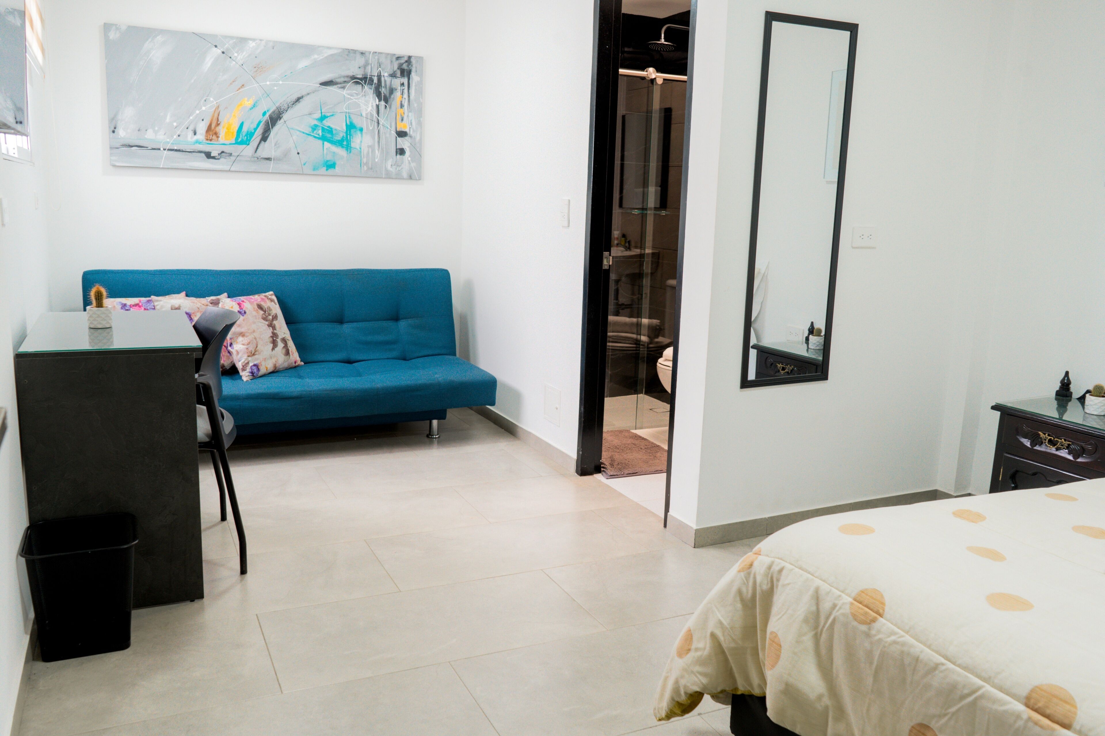 A modern bedroom with a blue sofa, a desk with a chair, a bed with a polka dot bedspread, and a large mirror.