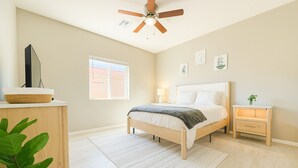 Een slaapkamer met een bed, nachtkastje, ventilator, raam en een plant.