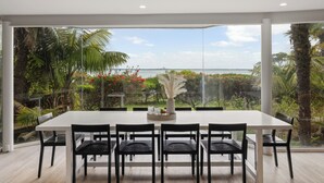 Un comedor con mesa blanca y sillas negras, ventanas amplias que dan a zonas verdes y una masa de agua.
