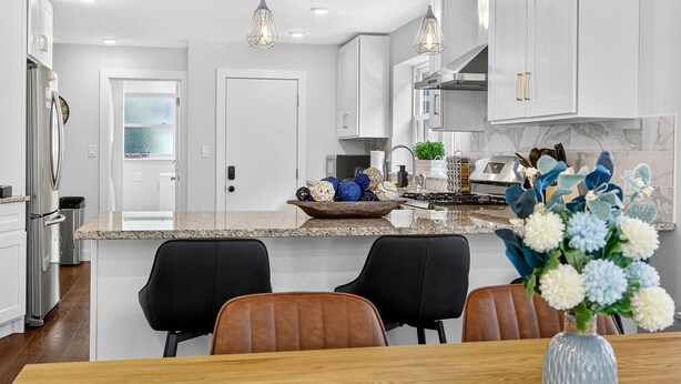 Een moderne keuken met witte kasten, granieten werkbladen en roestvrijstalen apparatuur. Er is een eethoek met een houten tafel en stoelen, en een vaas met bloemen op de tafel.