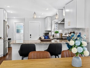 A modern kitchen with white cabinets, granite countertops, and stainless steel appliances. There is a dining area with a wooden table and chairs, and a vase with flowers on the table. - Hot Tub 3BR Somerville: Parking, Fire Pit, Near T (Somerville)