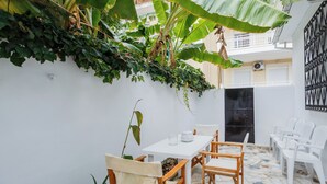 Un petit espace extérieur aménagé pour prendre les repas, avec une table et des chaises blanches, entouré d’un mur blanc et d’un grand bananier.