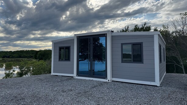 A small, single-story cabin with a sliding glass door and a window, situated on a gravel surface with a lake and trees in the background. - TH2-Enjoy the serenity of the lake in the comfort of this 2 bedroom tiny home. (Bridgeport)