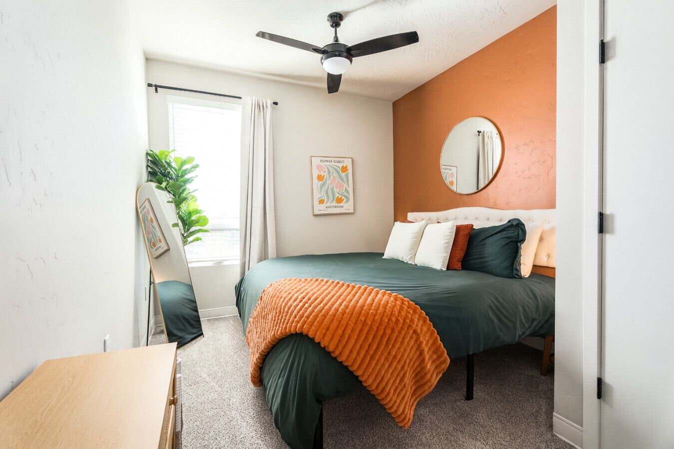A bedroom with a bed, a ceiling fan, a window with curtains, a plant, and a framed picture on the wall.