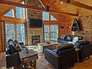 L’intérieur chaleureux d’un chalet avec une cheminée en pierre, des murs en bois et de grandes fenêtres offrant une vue sur la forêt environnante.