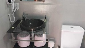 Une petite salle de bain avec une étagère en verre où sont posées des serviettes, un lavabo noir, des toilettes blanches et un sèche-cheveux branché sur une prise murale.
