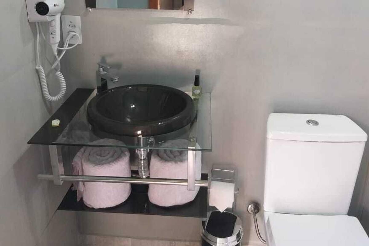 A compact bathroom with a glass shelf holding towels, a black sink, a white toilet, and a hairdryer on a wall socket.