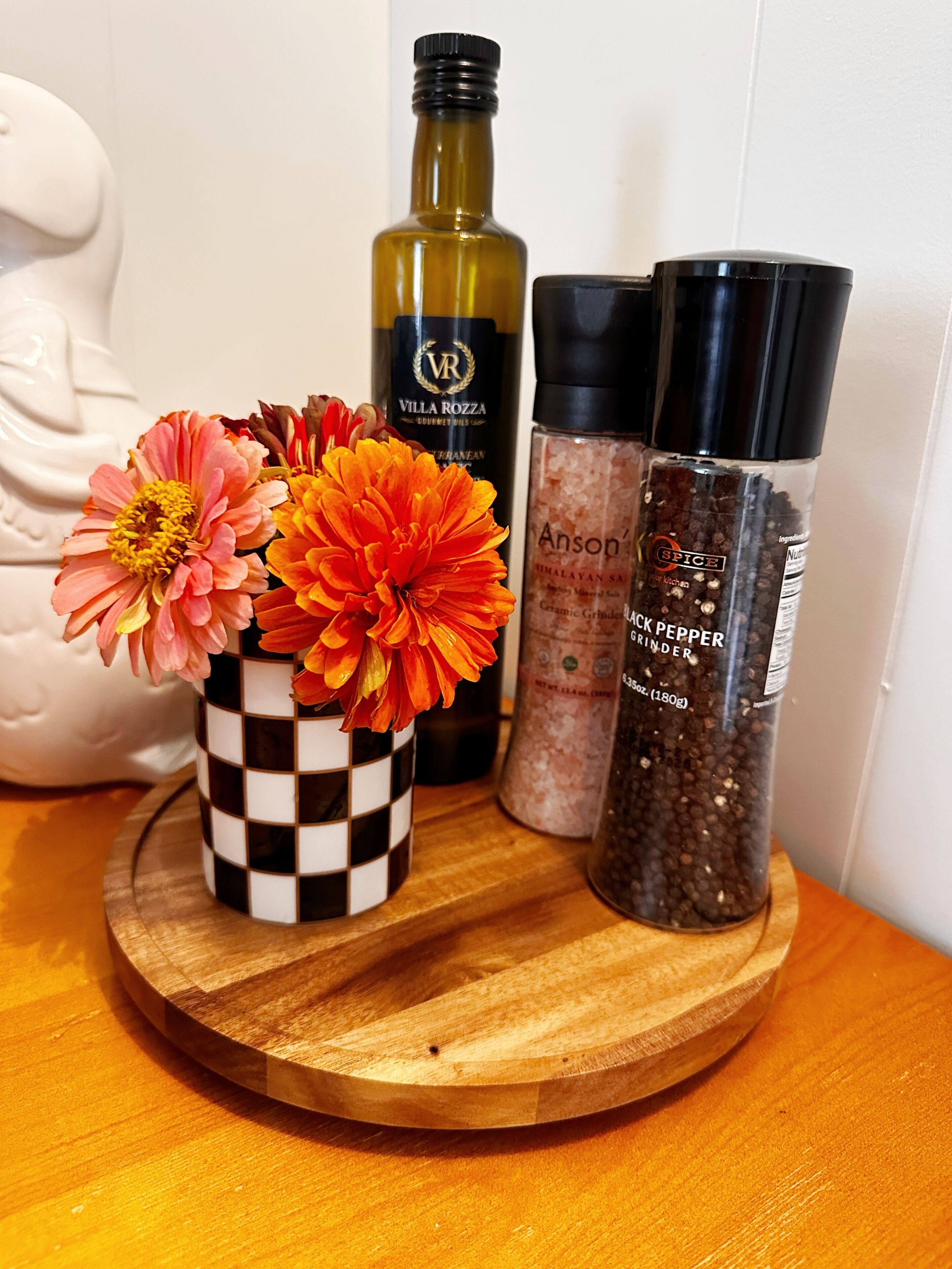 Un plateau en bois sur lequel se trouve un vase à carreaux contenant des fleurs, une bouteille d’huiles gastronomiques Villa Rozza, une bouteille de sel de l’Himalaya Anson et un moulin à poivre.