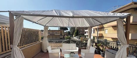 Un patio al aire libre con mobiliario de ratán blanco, una zona cubierta con cortinas blancas y plantas en macetas.