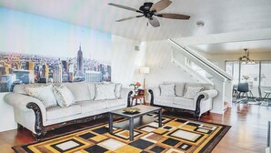 Ein Wohnzimmer mit Sofa, Couchtisch, Teppich mit Muster, Deckenventilator und einem Stadtbild an der Wand.