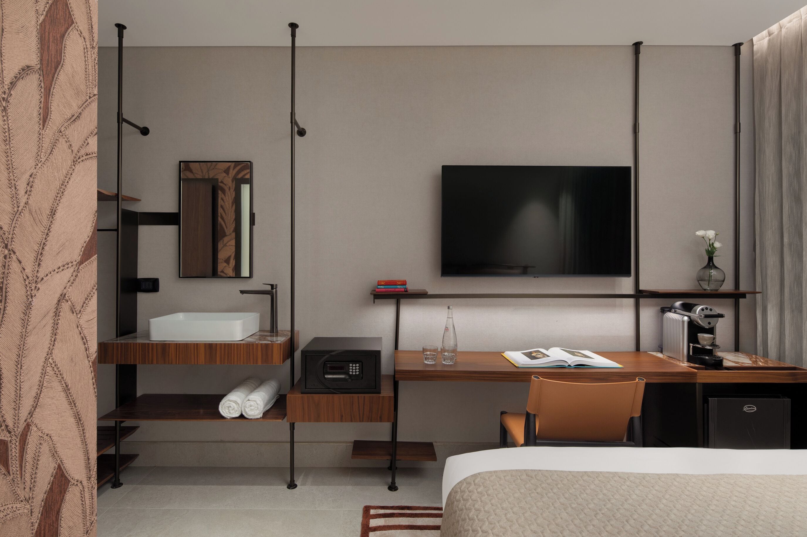 A hotel room with a bed, a desk with a computer, a TV, a mirror, and a shelf with books and a vase.