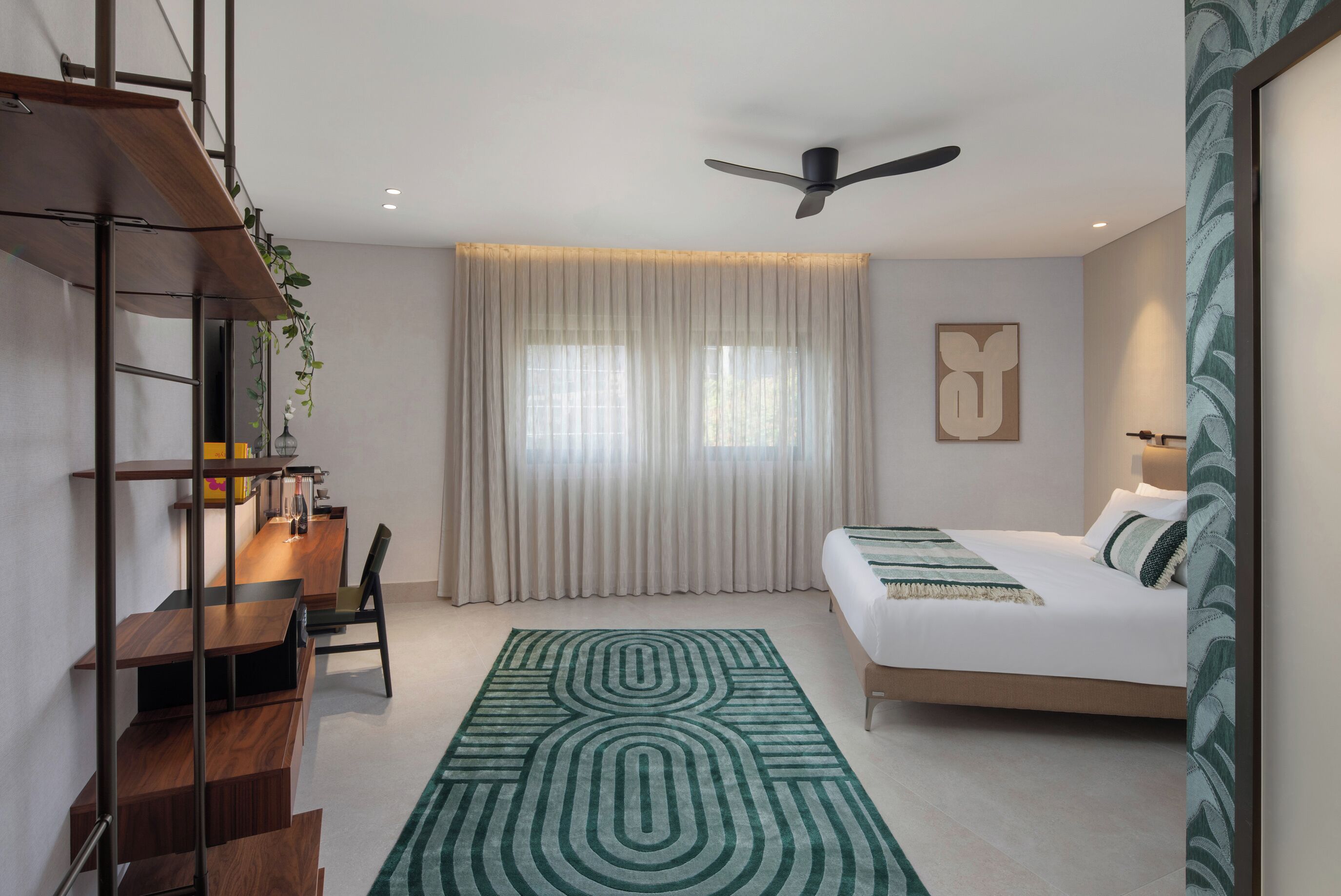 Una habitación de hotel moderna con un escritorio de madera, una cama con colcha verde y blanca estampada, un ventilador de techo y un ventanal con cortinas traslúcidas.