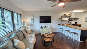 A living room with a sofa, a coffee table, and a TV. There is a kitchen area with a bar and stools. - 30-A Getaway near Seaside, FL --Great for Snowbirds, couples, or small families (Santa Rosa Beach)