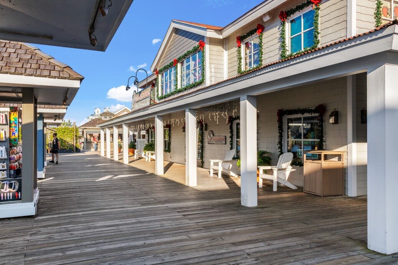A covered wooden deck with white chairs and tables, a store on the left, and a building with windows decorated with greenery and red ribbons.