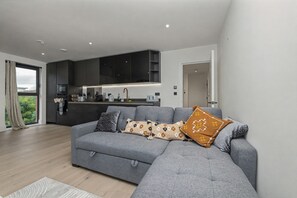 A modern living room with a grey sectional sofa, a kitchen area in the background, and a view through a large window. - Canada Water Suite | 1 bedroom flat | Rotherhithe, London (London)