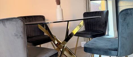 A dining area with a glass table, four chairs with gold legs, and a framed artwork on the wall.