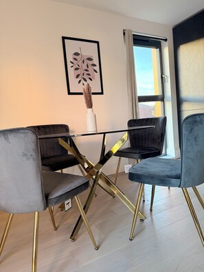 Uma sala de jantar com mesa de vidro, quatro cadeiras com pés dourados e uma obra de arte emoldurada na parede.