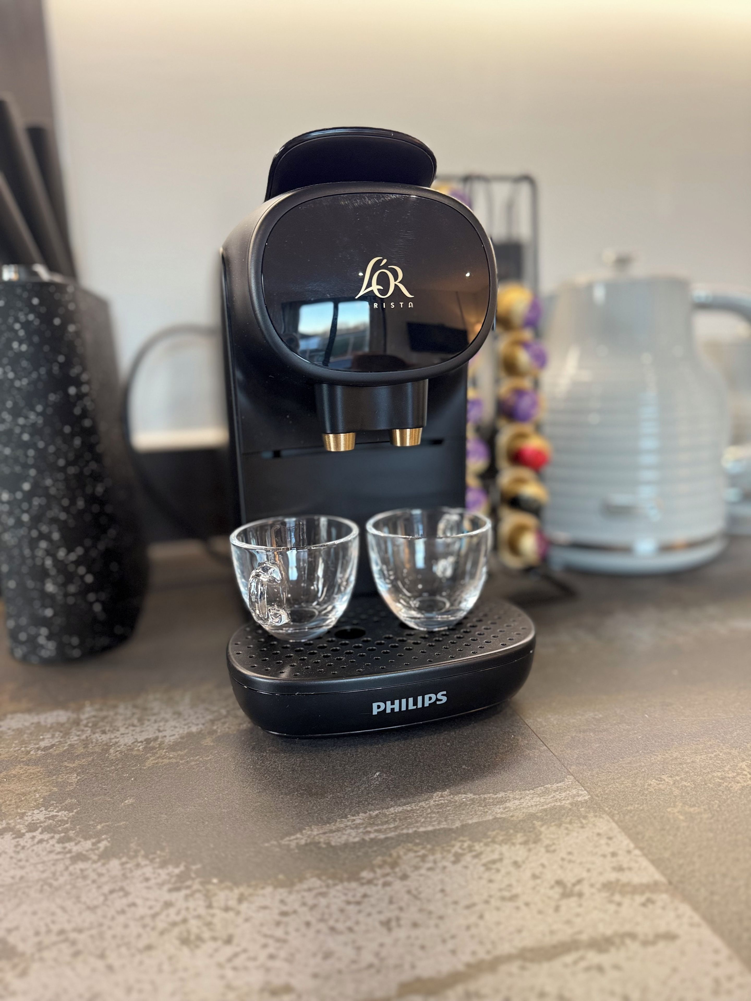 A coffee maker with two cups placed on its base, a kettle, and a decorative vase in the background.