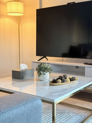 A modern living room with a flat-screen TV, a white marble coffee table, a grey sofa, a floor lamp, a white vase with green plants, and a bowl of decorative balls. - Canada Water Suite | 1 bedroom flat | Rotherhithe, London (London)