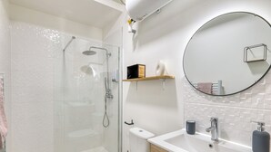 A modern bathroom with a glass-enclosed shower, a toilet, a sink with a mirror, and a shelf with toiletries. - Charming air-conditioned T2 near Lyon and the metro (Villeurbanne)
