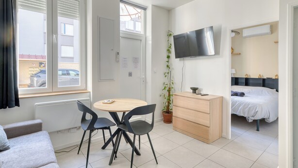 A compact room with a bed, a TV, a dining area with chairs, a small table, a potted plant, and a window with blinds. - Charming air-conditioned T2 near Lyon and the metro (Villeurbanne)