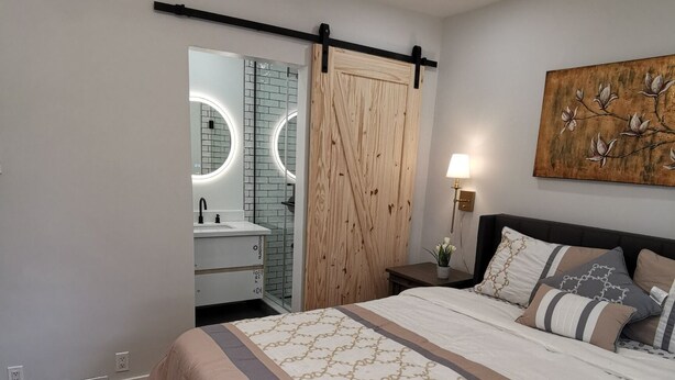 Een slaapkamer met een groot bed, een houten garderobe, een badkamer met een spiegel en ronde verlichting, en een kunstwerk aan de wand.