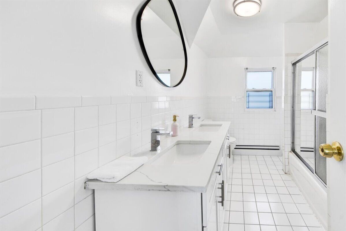 Un baño moderno con azulejos blancos, una encimera alargada con un lavabo, un espejo y una ventana.