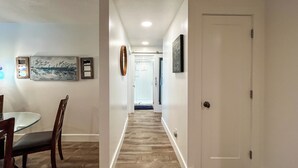 A hallway with a dining area on the left, featuring a table and chairs. The hallway has a wooden floor and white walls with framed artwork. A door is visible on the right side. - Extended Ski/Work/Visit secluded Cul-da-sac cutie (Salt Lake City)