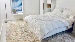 Un dormitorio con una cama grande, una mesita de noche, una lámpara en la mesita, un cuadro enmarcado en la pared y una estantería.