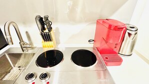 Una cocina con un lavamanos de acero inoxidable, dos hornillas negras, una cafetera roja y un hervidor plateado.