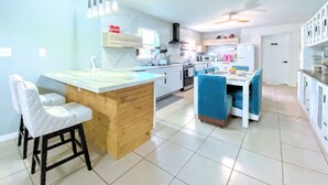 Cocina moderna con una isla central, taburetes de bar, una zona de comedor con sillas azules y un refrigerador blanco.