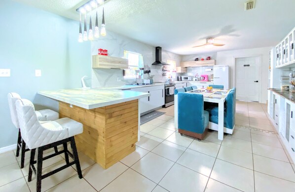 A modern kitchen with a central island, bar stools, a dining area with blue chairs, and a white refrigerator.