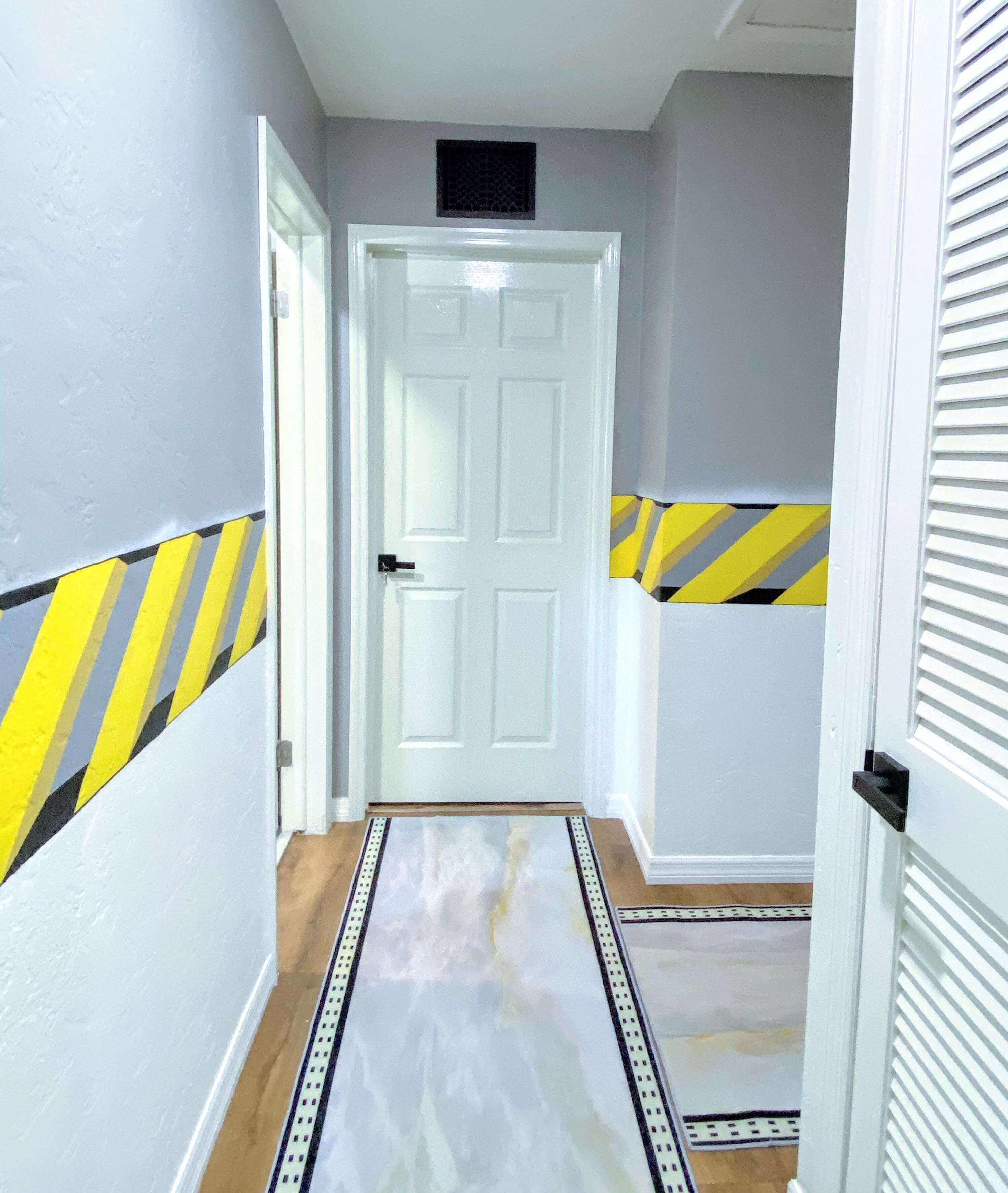 Un pasillo con una puerta blanca, cinta de precaución amarilla y negra en las paredes y un tapete estampado en el suelo.