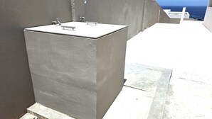 A grey, rectangular outdoor water feature with a faucet and a metal handle, adjacent to a building with a concrete exterior and a view of the sea. - Romantic Studio: Ocean, Salt Pans with Flamingos & Infinity Pool (Willemstad)