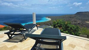 A terrace with two lounge chairs and a table, overlooking a coastal landscape with a clear view of the ocean and a hilly terrain. - Romantic Studio: Ocean, Salt Pans with Flamingos & Infinity Pool (Willemstad)