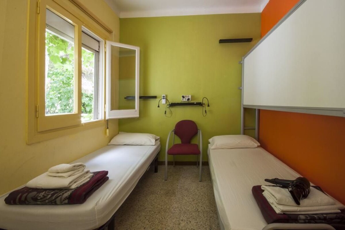 A small, single-room accommodation with two single beds, a chair, and a window.