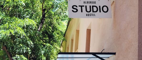 A sign for 'Albergue Studio Hostel' with a view of a tree and a building facade.