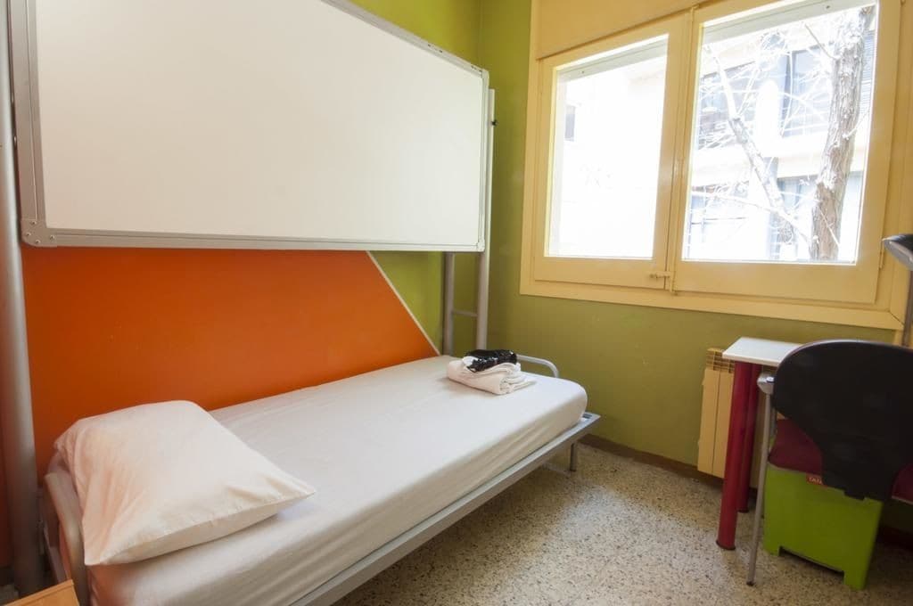 Un lit simple avec un oreiller blanc, un tableau blanc au-dessus, une fenêtre donnant sur des arbres et un petit bureau avec une chaise.