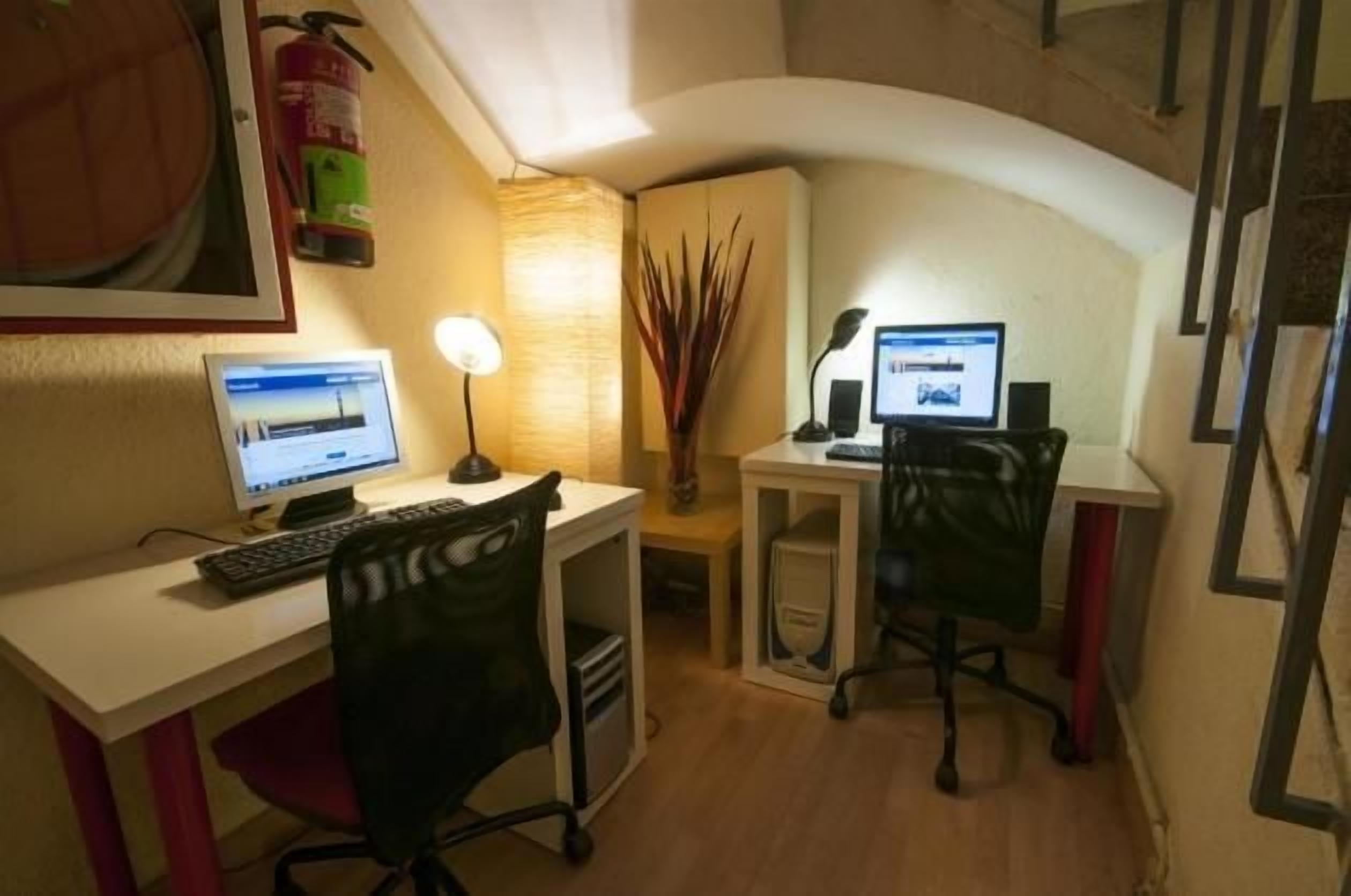 A small office with two desks, each with a computer and a lamp. There is a fire extinguisher on the wall and a plant in the corner.