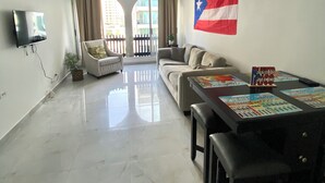 Ein geräumiges Wohnzimmer mit einer großen puertoricanischen Flagge, einem Deckenventilator, einem Flachbildfernseher, einem Esstisch mit Unterlagen und Blick durch ein großes Fenster.