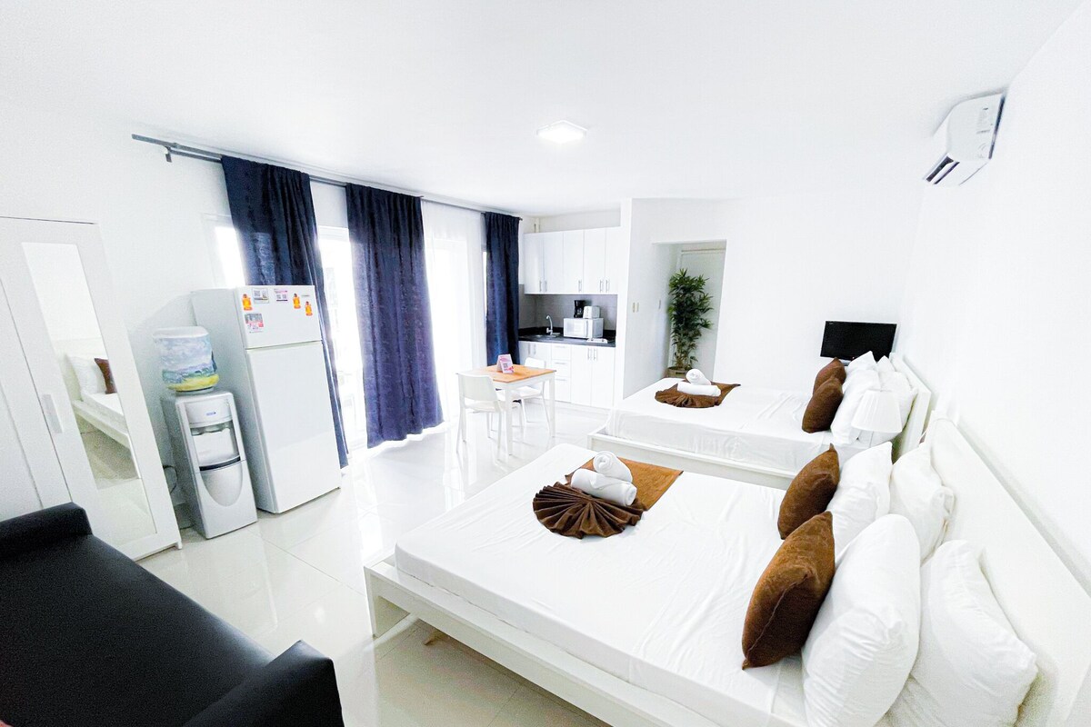 A hotel room with a bed, a desk, a chair, a small kitchen area, and a bathroom.