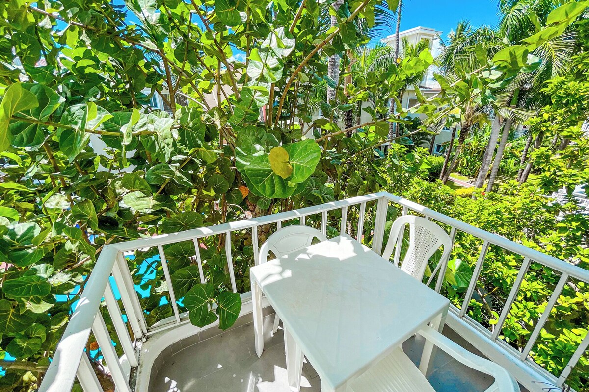 A balcony with white chairs and a table, surrounded by lush green foliage and palm trees.