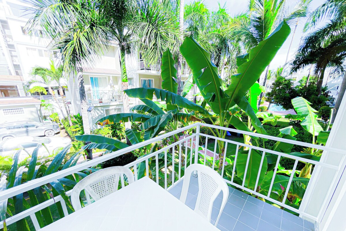 A balcony with a white table and chairs, surrounded by lush green plants and a view of a residential area.