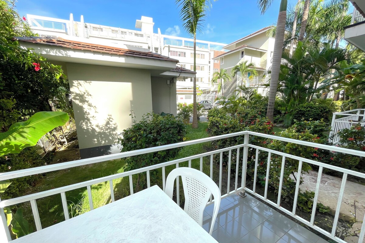 A balcony with a white table and chair, surrounded by lush greenery and a view of a building with a tiled roof.