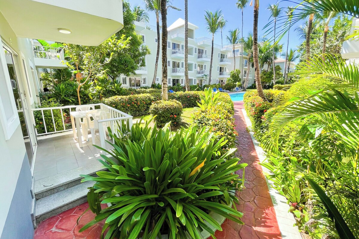 A hotel courtyard with a swimming pool, lush greenery, and a white building with balconies.