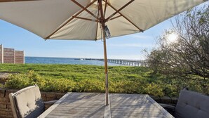 Eine Holzterrasse mit Tisch und Stühlen, einem Sonnenschirm, Blick auf das Meer und einem Pier im Hintergrund.