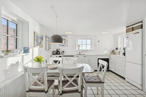 A modern kitchen with white cabinetry, a dining table with chairs, a pendant light, and a window with a view of trees. - Family-friendly beachside holiday home (Rønne)