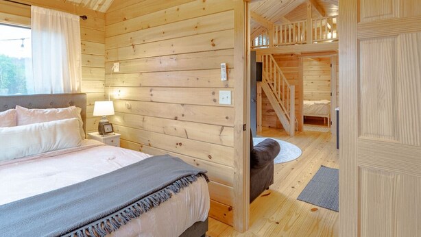 Une chambre à coucher dans une cabane en bois, avec un lit, une table de chevet et une vue sur l’escalier menant à l’étage supérieur.