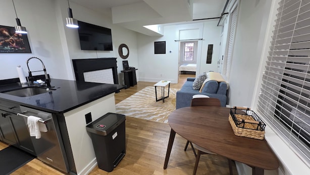 A modern kitchen with a black countertop, a sink, and a trash bin. There is a dining area with a wooden table and chairs. - Quiet cheerful condo in the heart of OTR! (Cincinnati)